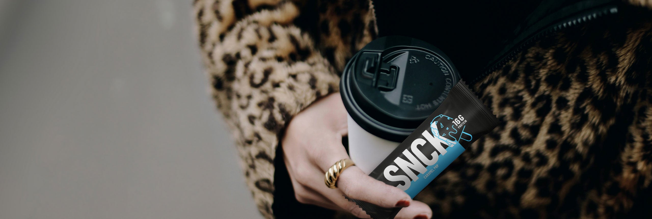 Person holding a coffee cup with a SNCK bar, wearing a leopard print coat.